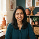 A professional portrait of a smiling South Asian woman in a modern home office with warm amber and teal decor.